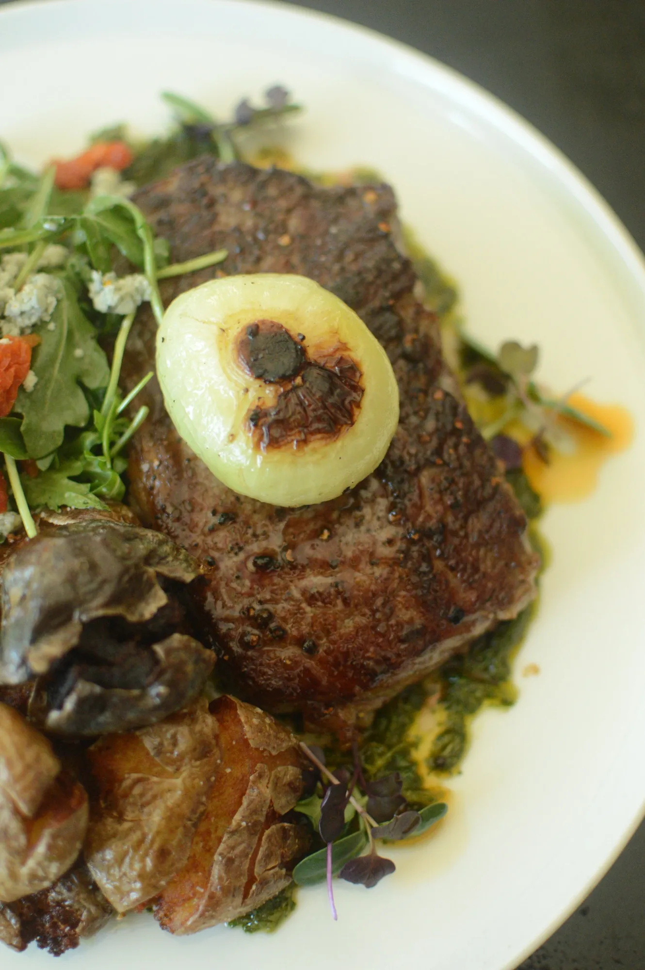 A strip steak crowned with an onion ring over greens and roasted vegetables.