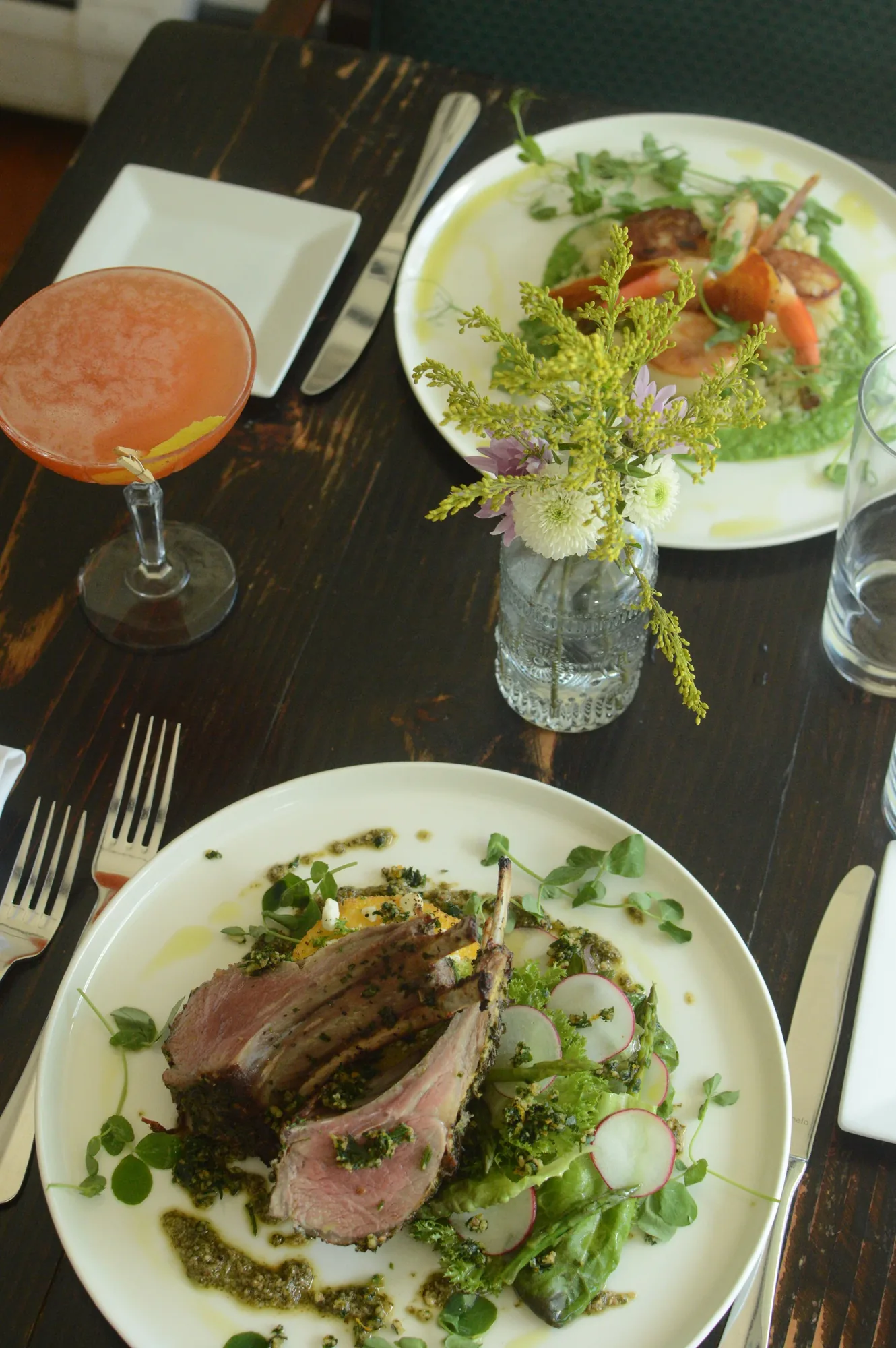 A dinner table set with a plated dish, a salad and a cocktail on dark wood.